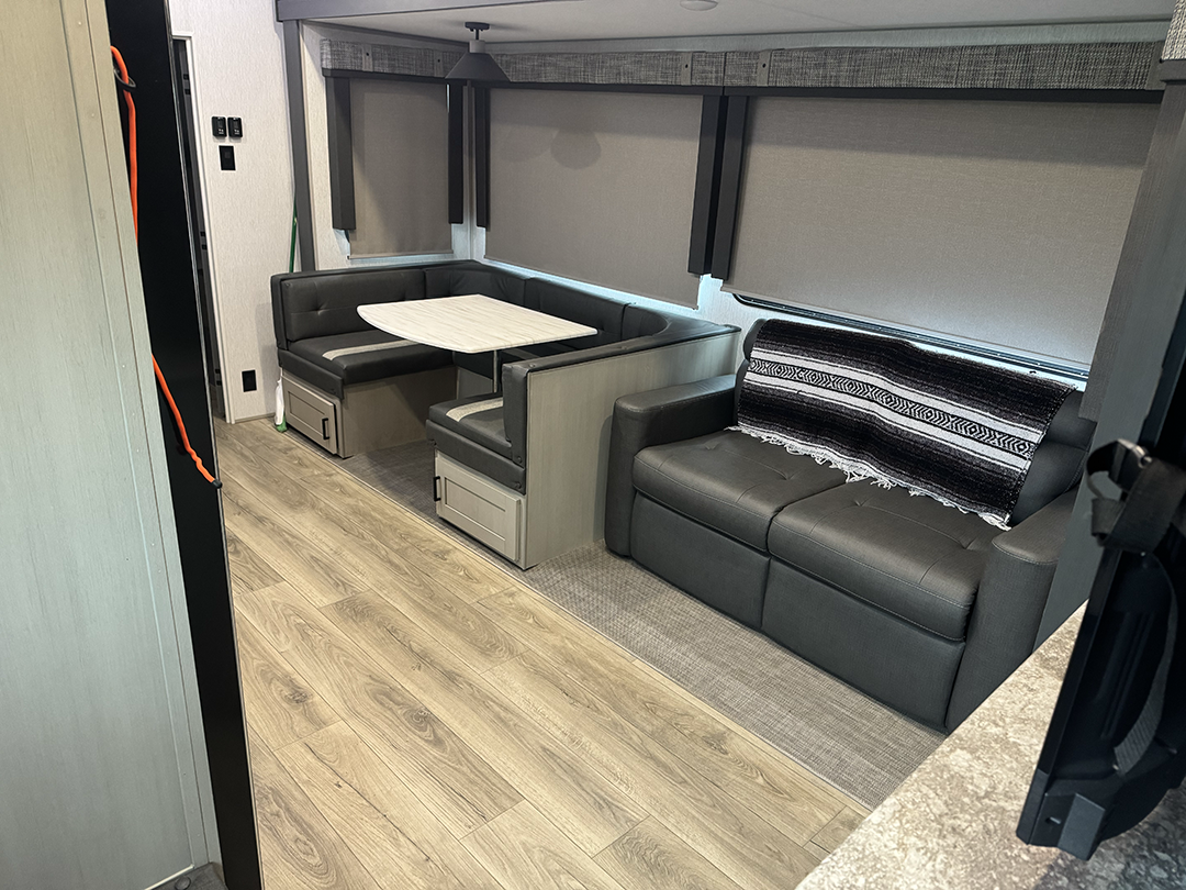 Interior of a modern RV living area with a gray L-shaped booth dining area and a matching sofa, a light wood table, and large windows with shades down.