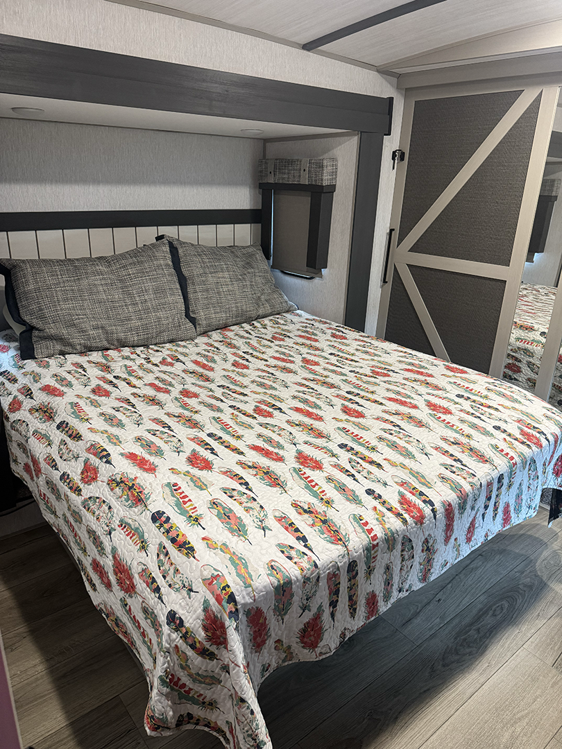 Queen bed in a modern RV bedroom with a colorful feather-pattern quilt and two gray pillows. A sliding barn-door closet is visible nearby.