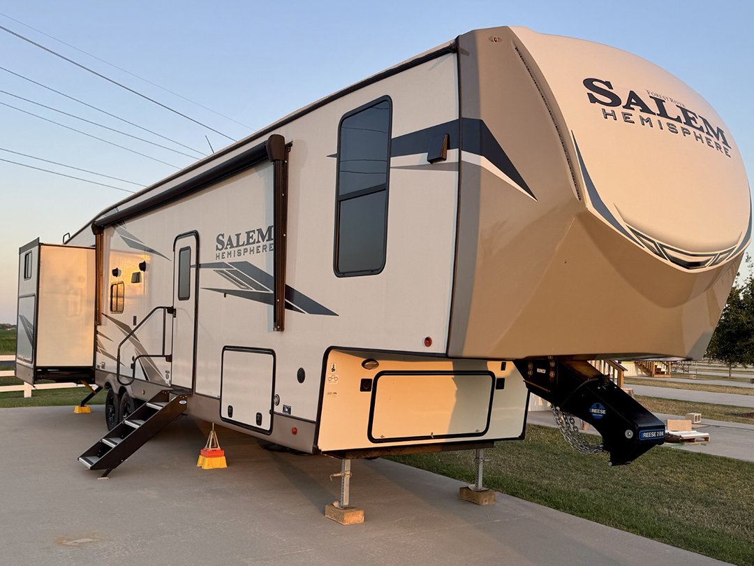 Side view of a beige and gray Salem Hemisphere fifth-wheel trailer with a slide-out extended and entry stairs down on a concrete pad at sunset