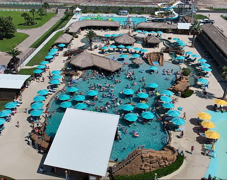 Aerial view of a crowded water park with bright turquoise umbrellas around a large pool and sandy cabanas, slides in the background.