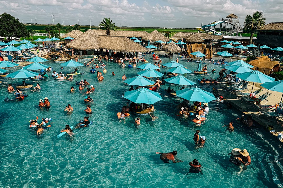 A crowded outdoor pool area with turquoise umbrellas, people swimming and lounging among thatched-roof huts and water slides in the background.